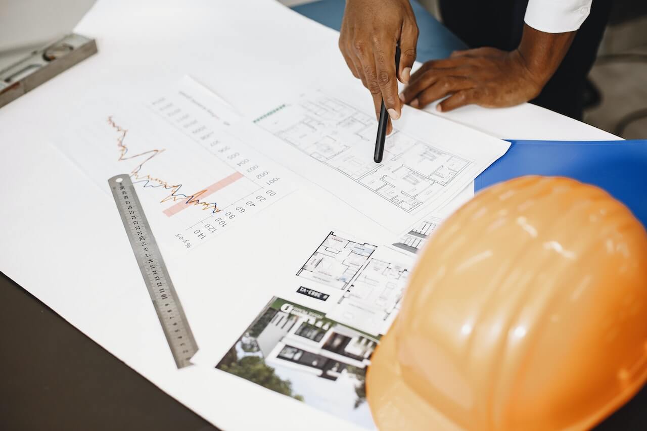 An engineer working on a drafting table holding a pencil pointing at floor plan drawing with other tools on the table like ruler and orange hard hat